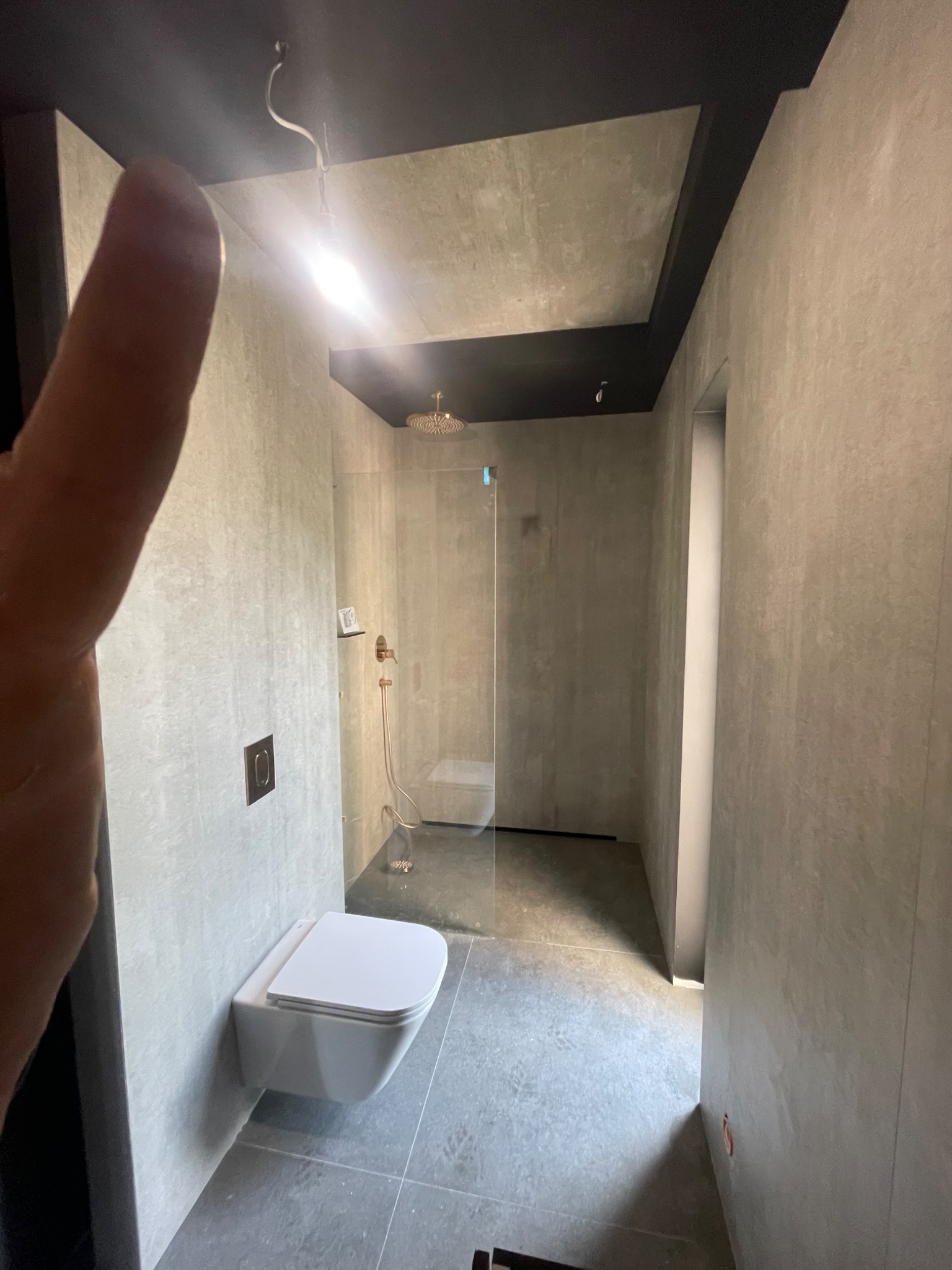 Nowoczesna łazienka z kabiną prysznicową typu walk-in, wiszącą toaletą i szarymi płytkami, oświetlona punktowym światłem, z widocznym fragmentem dłoni w kadrze.