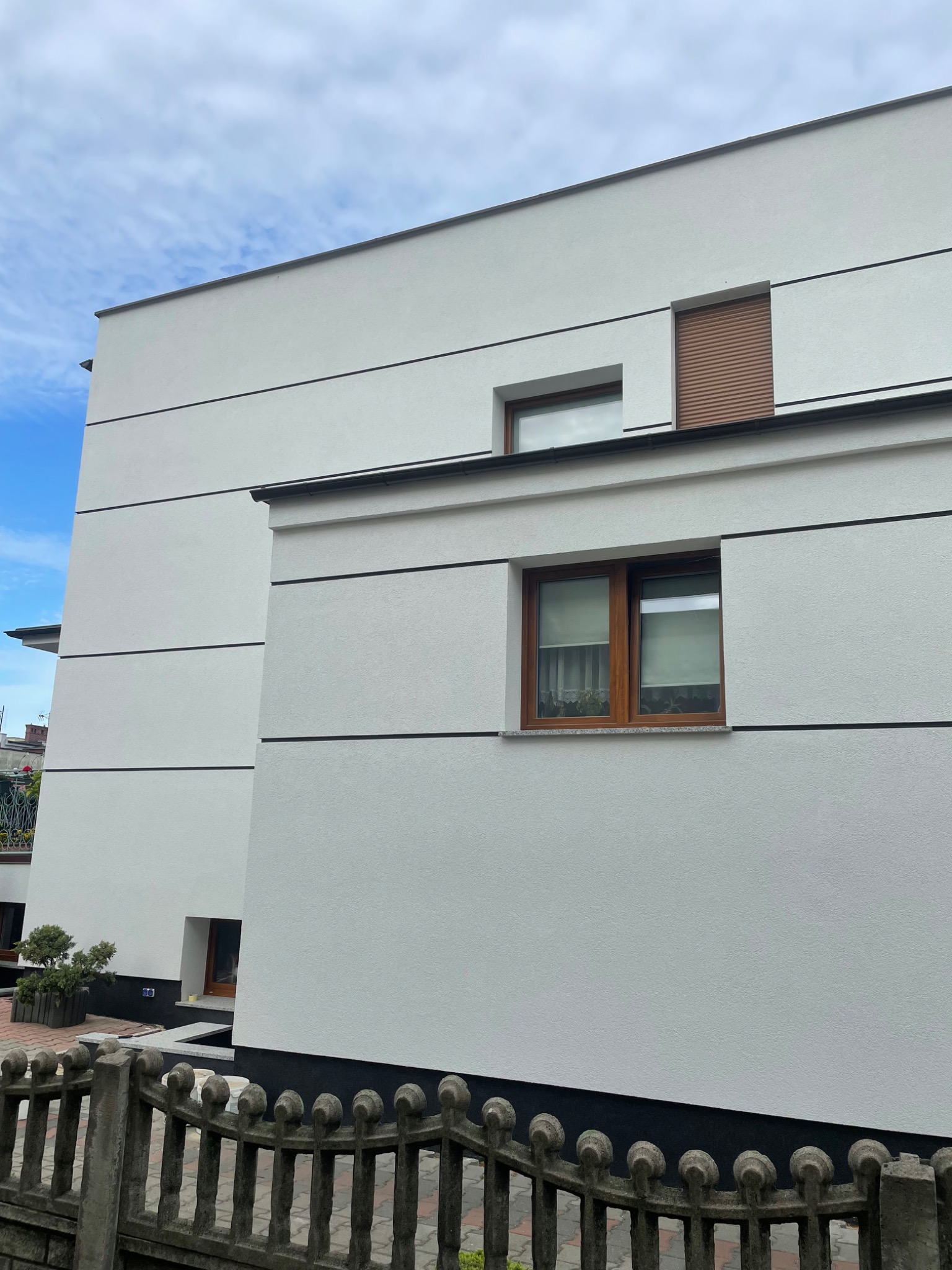 Nowoczesna, biała elewacja domu z poziomymi, czarnymi liniami dekoracyjnymi, drewnianą stolarką okienną i szarą, betonową podmurówką, widziana zza starego, betonowego płotu.