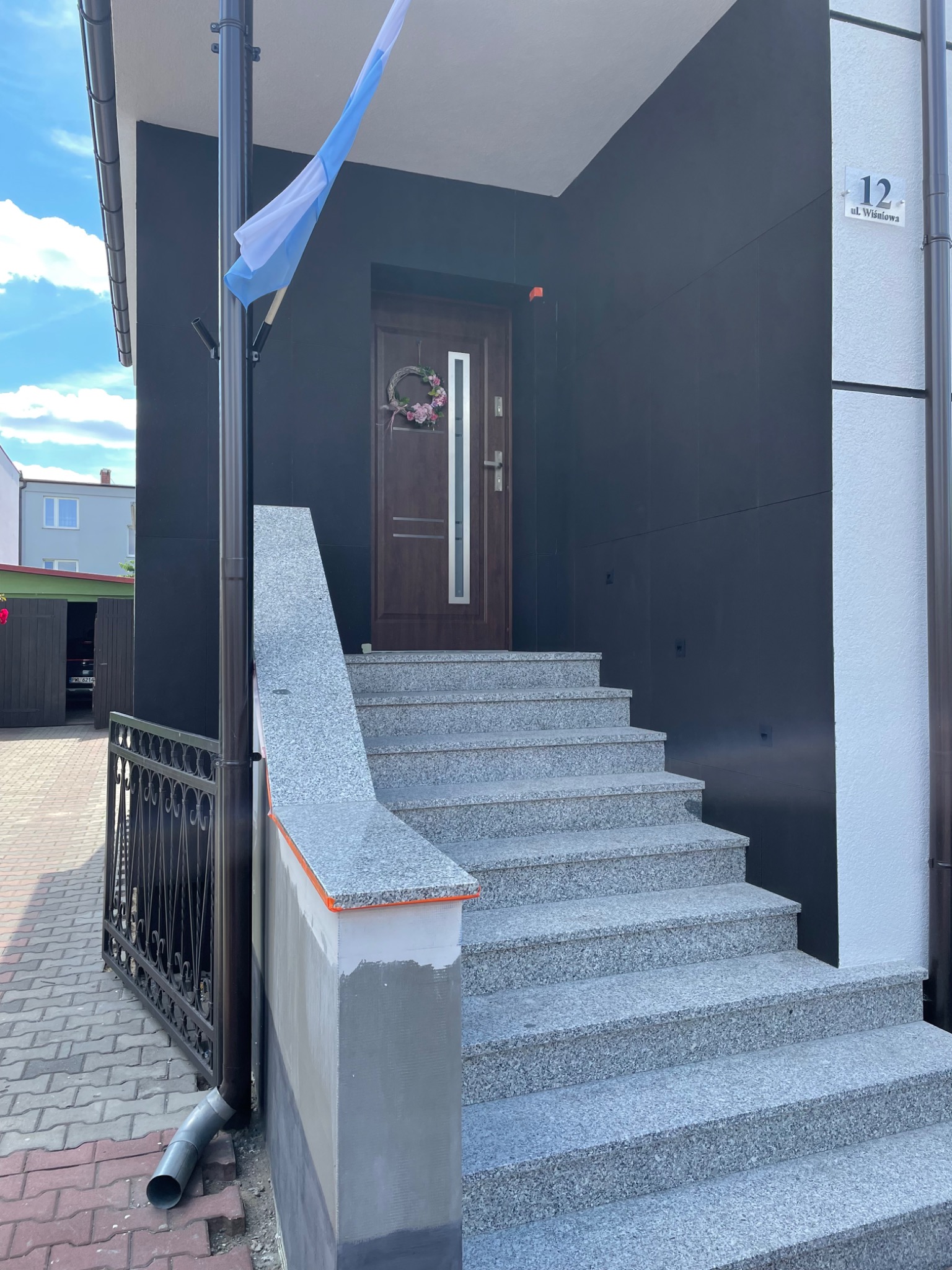 Wejście do domu z ciemną elewacją, granitowymi schodami i dekoracyjnym wieńcem na drzwiach. Widoczny fragment flagi i numer domu.