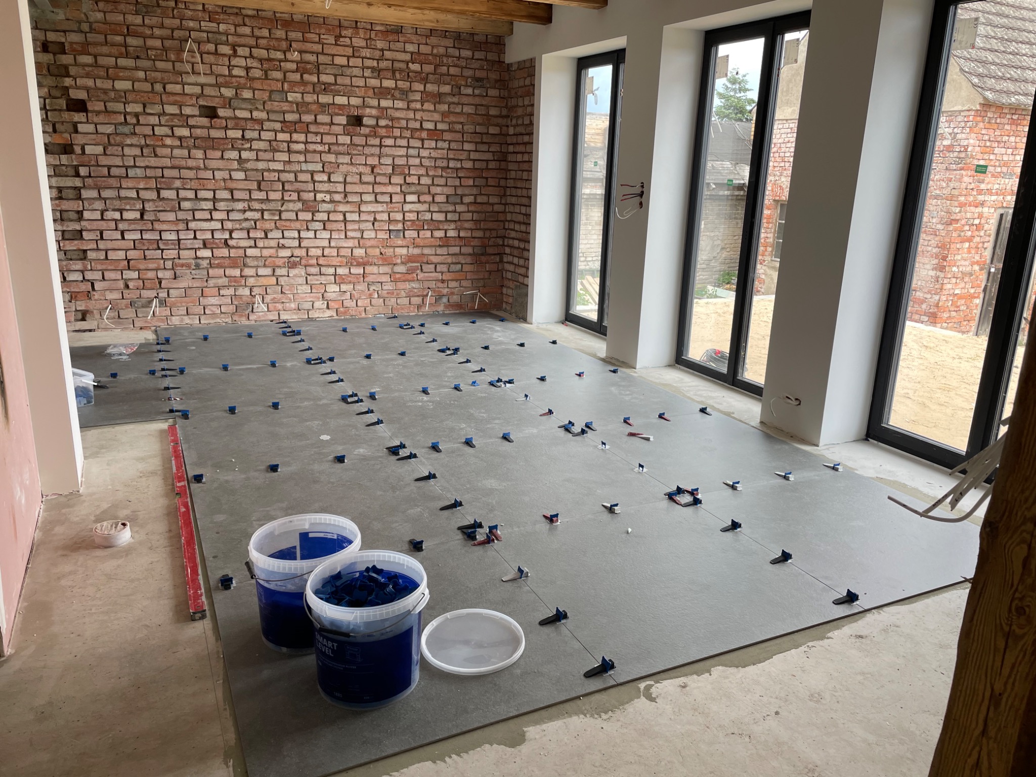 Układanie szarych płytek podłogowych z systemem poziomowania, widoczne dwa wiadra z elementami systemu, ceglana ściana i duże okna w tle.