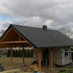 Nowy budynek gospodarczy z ciemną dachówką ceramiczną, drewnianą konstrukcją werandy i kominem, widoczny w trakcie budowy, z materiałami budowlanymi na pierwszym planie.