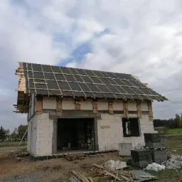 Budowa domu jednorodzinnego: widok na częściowo pokryty dach z czarnej dachówki, widoczne łaty, niezabudowane ściany z pustaków, teren budowy z materiałami budowlanymi.