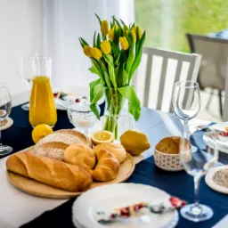 Stół nakryty do śniadania z pieczywem, cytrusami, dzbankiem soku pomarańczowego i bukietem żółtych tulipanów w wazonie, przygotowany do sesji home staging.