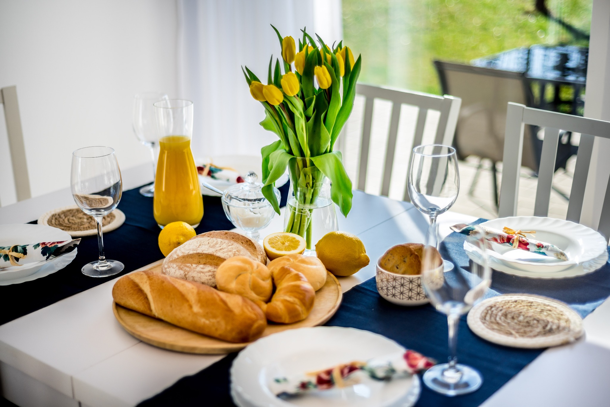 Stół nakryty do śniadania z pieczywem, cytrusami, dzbankiem soku pomarańczowego i bukietem żółtych tulipanów w wazonie, przygotowany do sesji home staging.