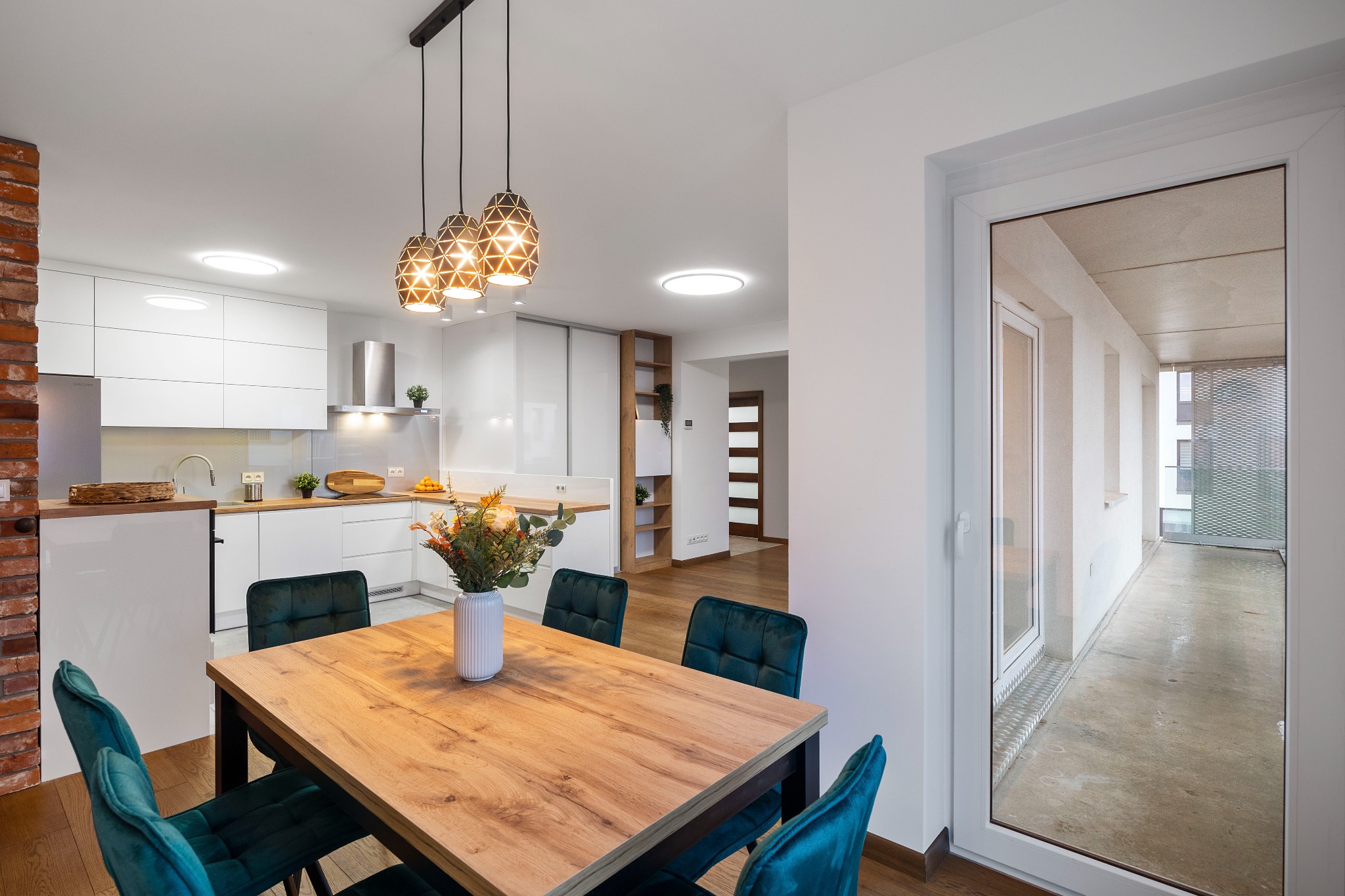 Jasna kuchnia z drewnianym stołem i turkusowymi krzesłami. W tle białe szafki i ceglana ściana. Widok na balkon przez przeszklone drzwi.