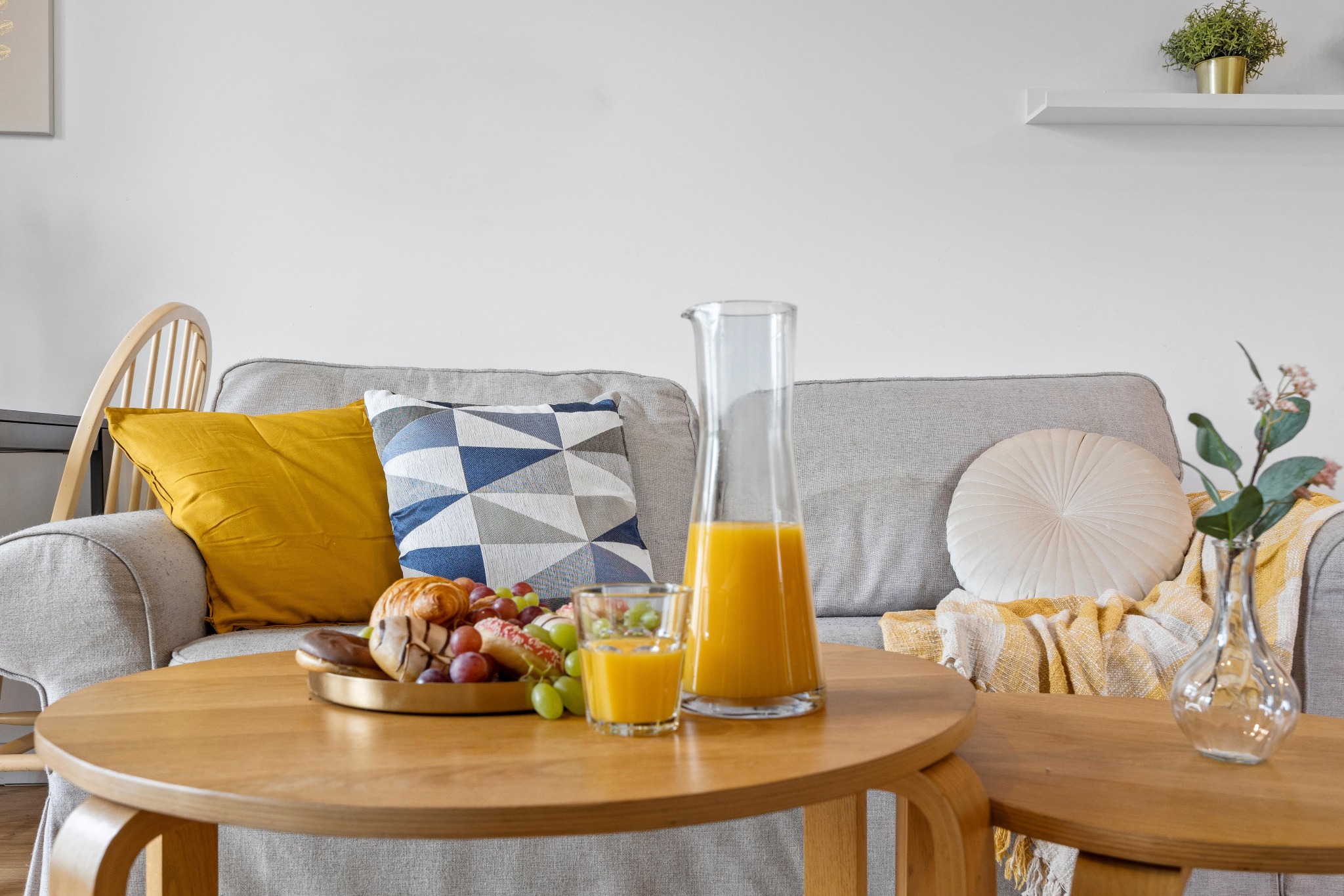 Jasne wnętrze: stoliki kawowe z przekąskami i sokiem, szara sofa z dekoracyjnymi poduszkami. Roślina w doniczce na ścianie. Przytulna aranżacja mieszkania.