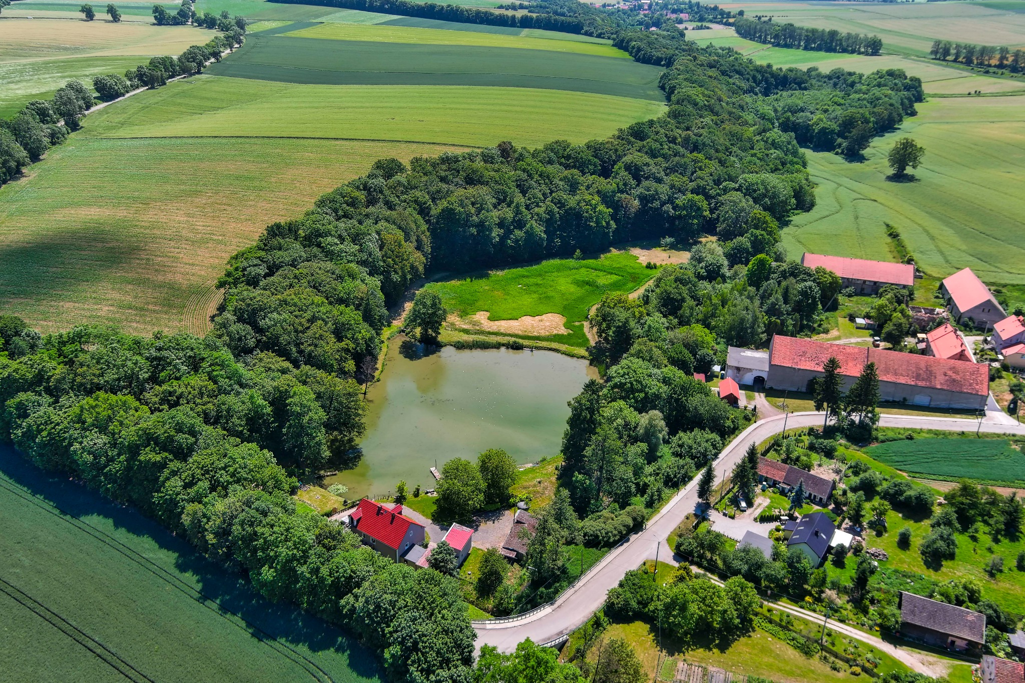 Panoramiczny widok z lotu ptaka: malownicze pola, lasy, staw i zabudowania wiejskie z czerwoną dachówką, tworzące krajobraz wiejski.