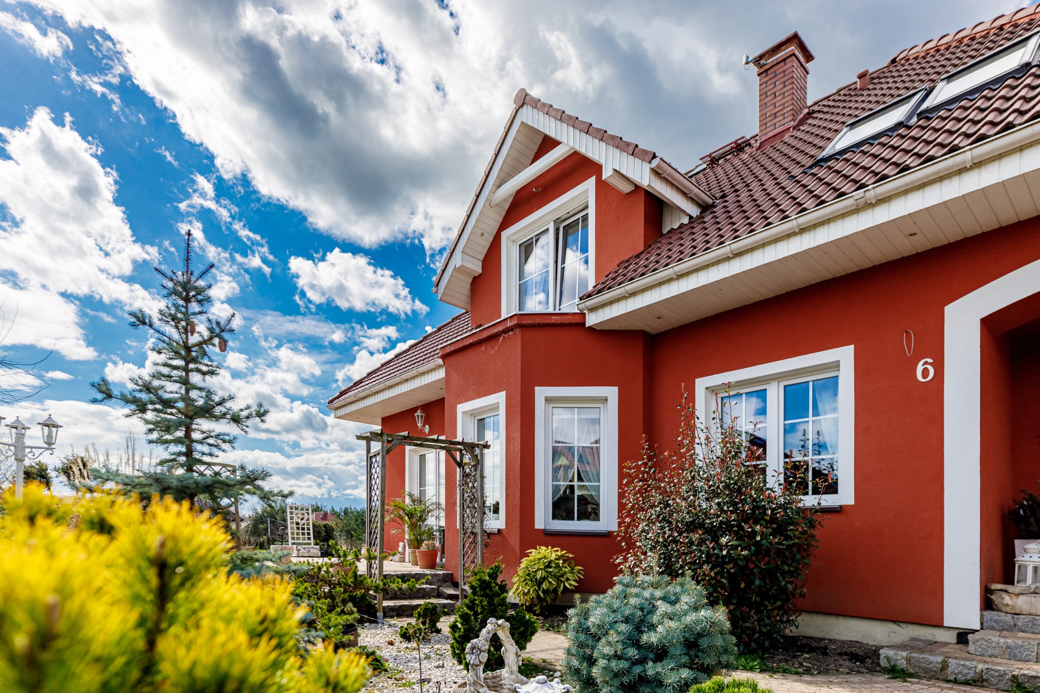 Czerwony dom jednorodzinny z białymi oknami i dachem z dachówki w Opolu, otoczony zielenią i niskimi krzewami, z numerem '6' widocznym na ścianie.