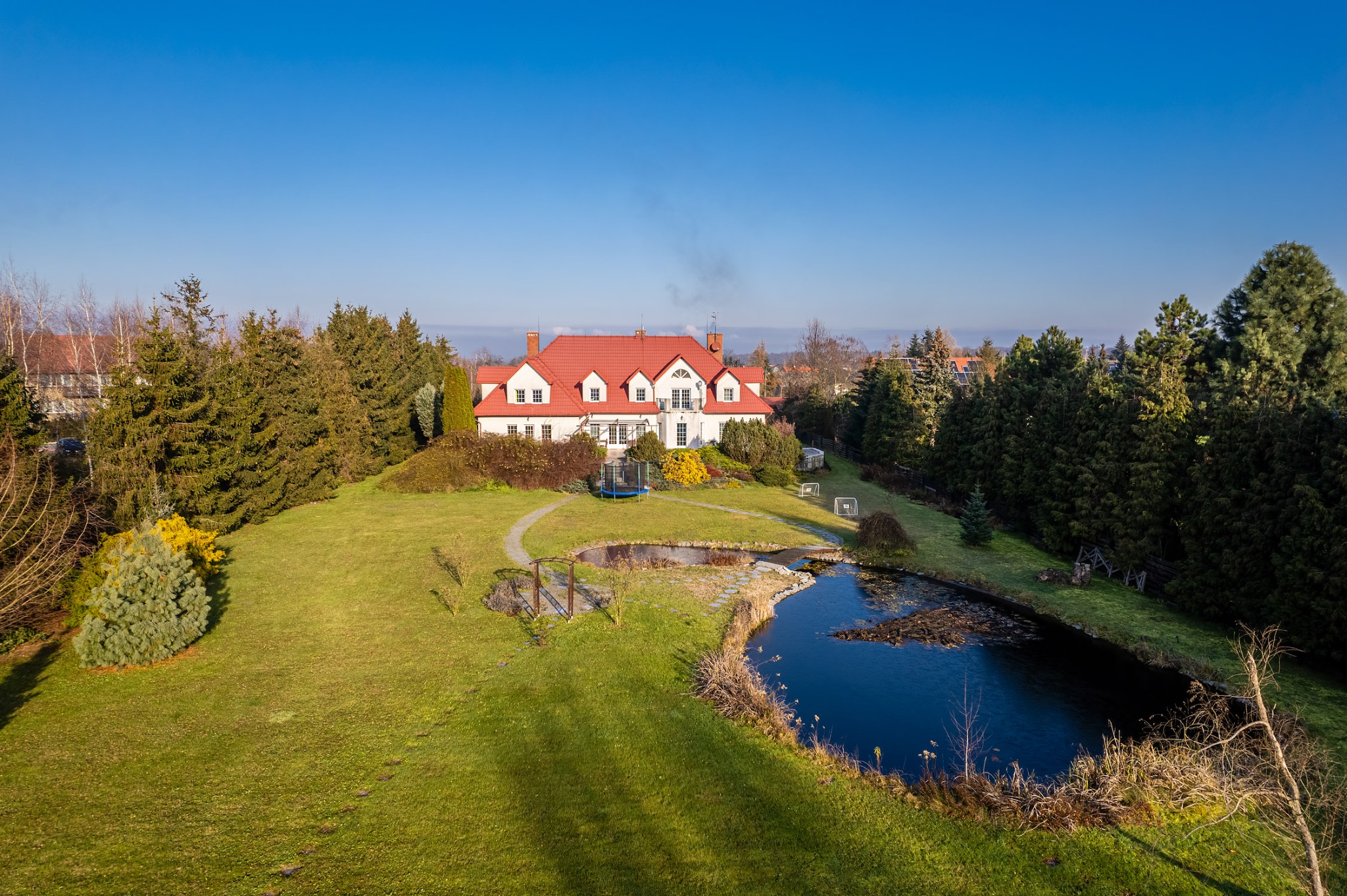 Panoramiczny widok z drona na duży, biały dom z czerwoną dachówką, otoczony zielonym trawnikiem, drzewami i stawem w słoneczny dzień. Na trawniku widoczny trampolina i małe bramki do piłki nożnej.