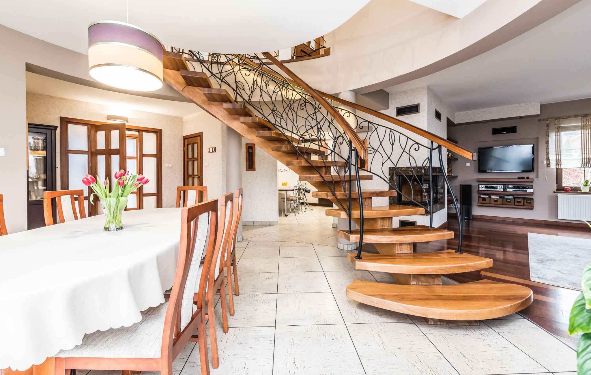 Przestronny salon z drewnianymi schodami spiralnymi z czarną, ozdobną balustradą, białym stołem z bukietem tulipanów i telewizorem na ścianie