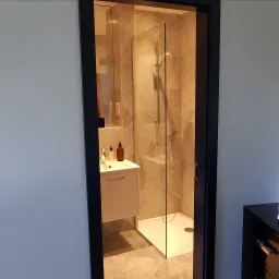 Wykończona łazienka z kabiną prysznicową za szklaną ścianką, umywalką nablatową z szafką, marmurowymi płytkami na ścianach i podłodze oraz czarną ramą drzwi.