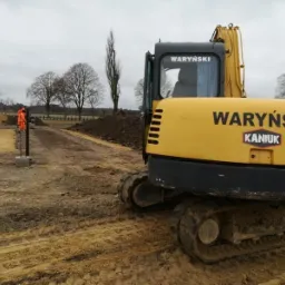 Żółta koparka firmy WARYŃSKI KANIUK na placu budowy z widocznymi fundamentami i pracownikami w pomarańczowych kombinezonach, pochmurne niebo w tle.