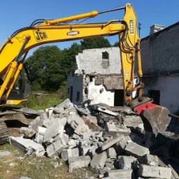Żółta koparka JCB z hydraulicznymi nożycami do betonu, rozbiórka częściowo zburzonego budynku z cegły i betonu, gruz na pierwszym planie, widok z boku w słoneczny dzień.