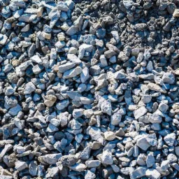 Góra szaro-białego kruszywa o zróżnicowanej granulacji, od drobnego pyłu po większe kamienie, oświetlona naturalnym światłem słonecznym.