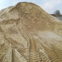 Duża pryzma piasku z wyraźnymi śladami opon pojazdu budowlanego, widoczny fragment betonowego muru w tle.