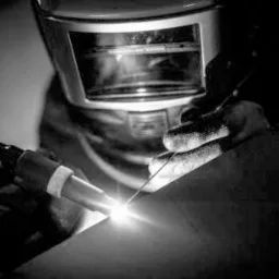 Spawacz w masce ochronnej wykonuje precyzyjne spawanie metodą TIG, widoczny intensywny blask łuku elektrycznego i elektroda wolframowa.