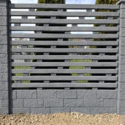Szary, betonowy płot z poziomymi panelami i słupkami, widok z bliska na tle zieleni i drzew