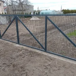 Nowo zamontowana, ciemnoszara brama panelowa z ukośnymi wzmocnieniami na betonowej podmurówce, widok z bliska na tle ogrodu z tunelem foliowym i budynkiem mieszkalnym.