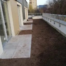 Nowoczesny balkon z przygotowanym podłożem pod trawnik i ułożonymi płytami chodnikowymi, widok na budynki w tle.