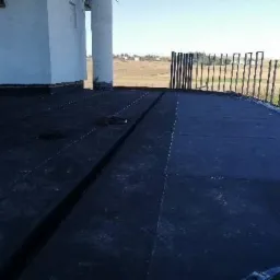 Czarna, płaska powierzchnia dachu pokryta papą, z widocznym systemem odprowadzania wody i częściowo zamontowaną balustradą w tle, w oddali krajobraz wiejski.