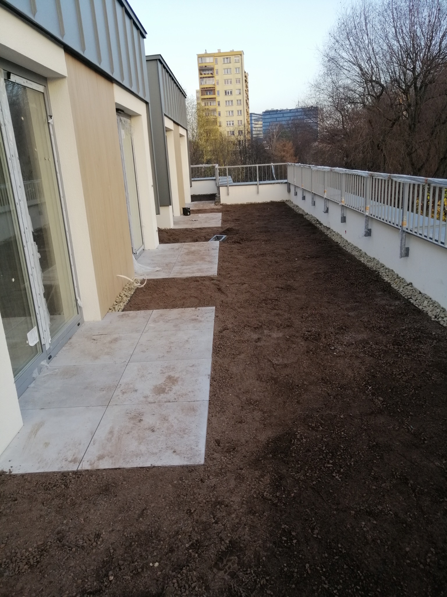 Nowoczesny balkon z przygotowanym podłożem pod trawnik i ułożonymi płytami chodnikowymi, widok na budynki w tle.