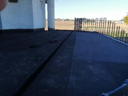 Czarna, płaska powierzchnia dachu pokryta papą, z widocznym systemem odprowadzania wody i częściowo zamontowaną balustradą w tle, w oddali krajobraz wiejski.