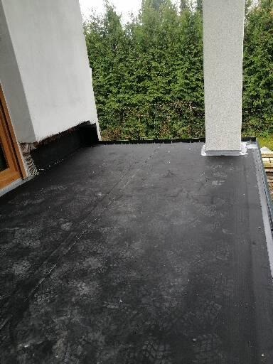 Czarna, płaska powierzchnia dachu pokryta papą, z widocznymi śladami butów i elementami konstrukcyjnymi, przylegająca do białej ściany budynku i kolumny, na tle zielonych krzewów.