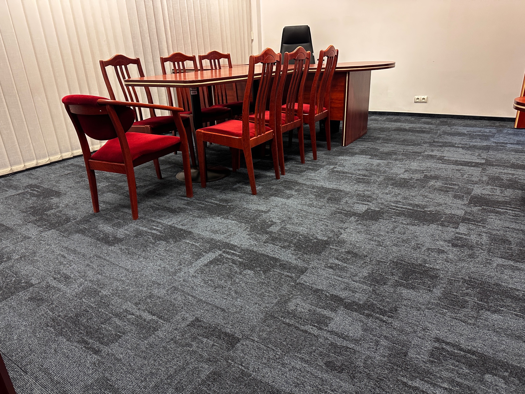 Biuro z szarą wykładziną dywanową w geometryczny wzór, mahoniowy stół konferencyjny z ośmioma czerwonymi krzesłami i biurko w tle.