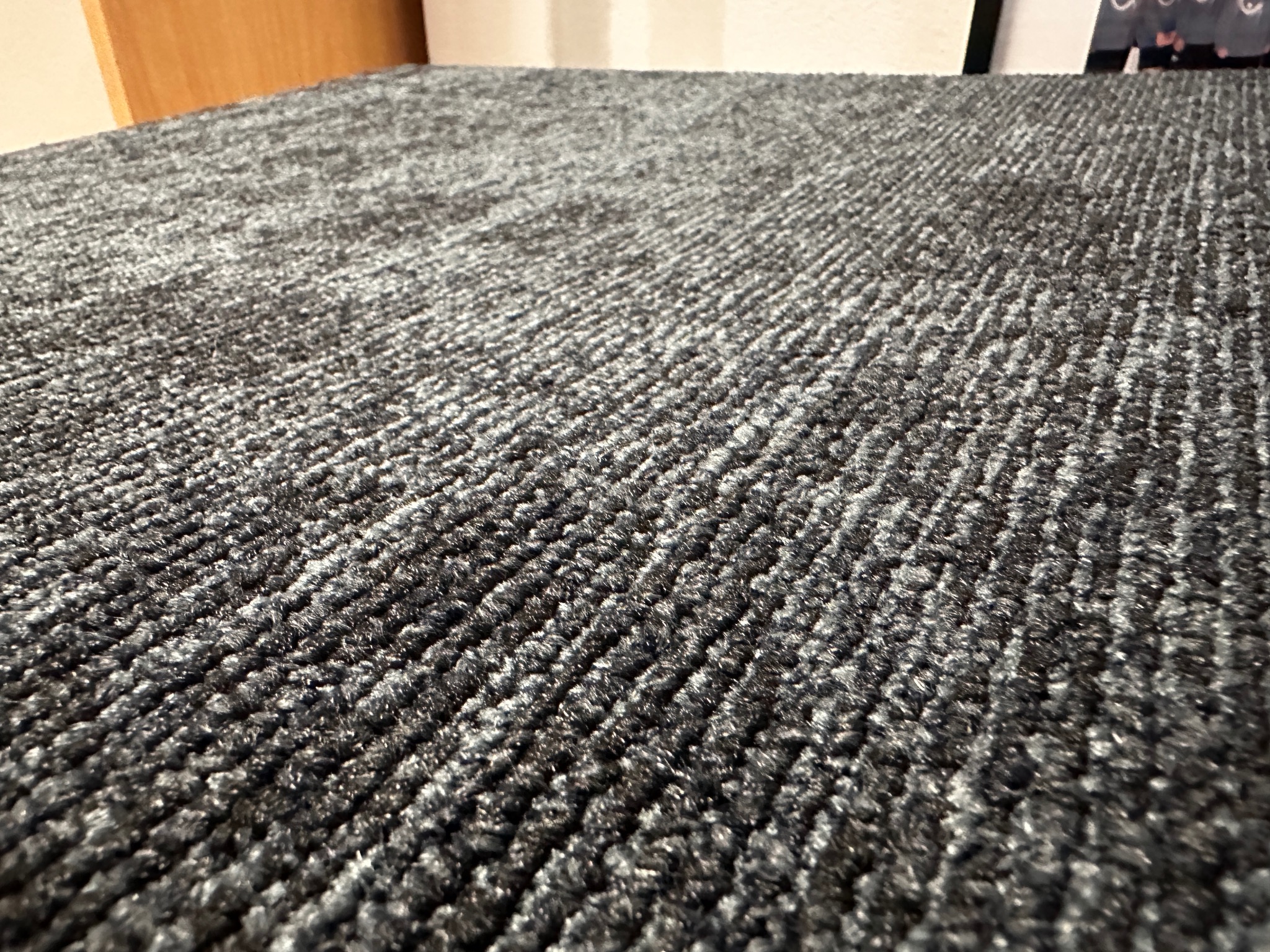 Szczegółowe ujęcie szarej wykładziny dywanowej o wyraźnej strukturze pętelkowej, ułożonej w pomieszczeniu z fragmentem mebla w tle.