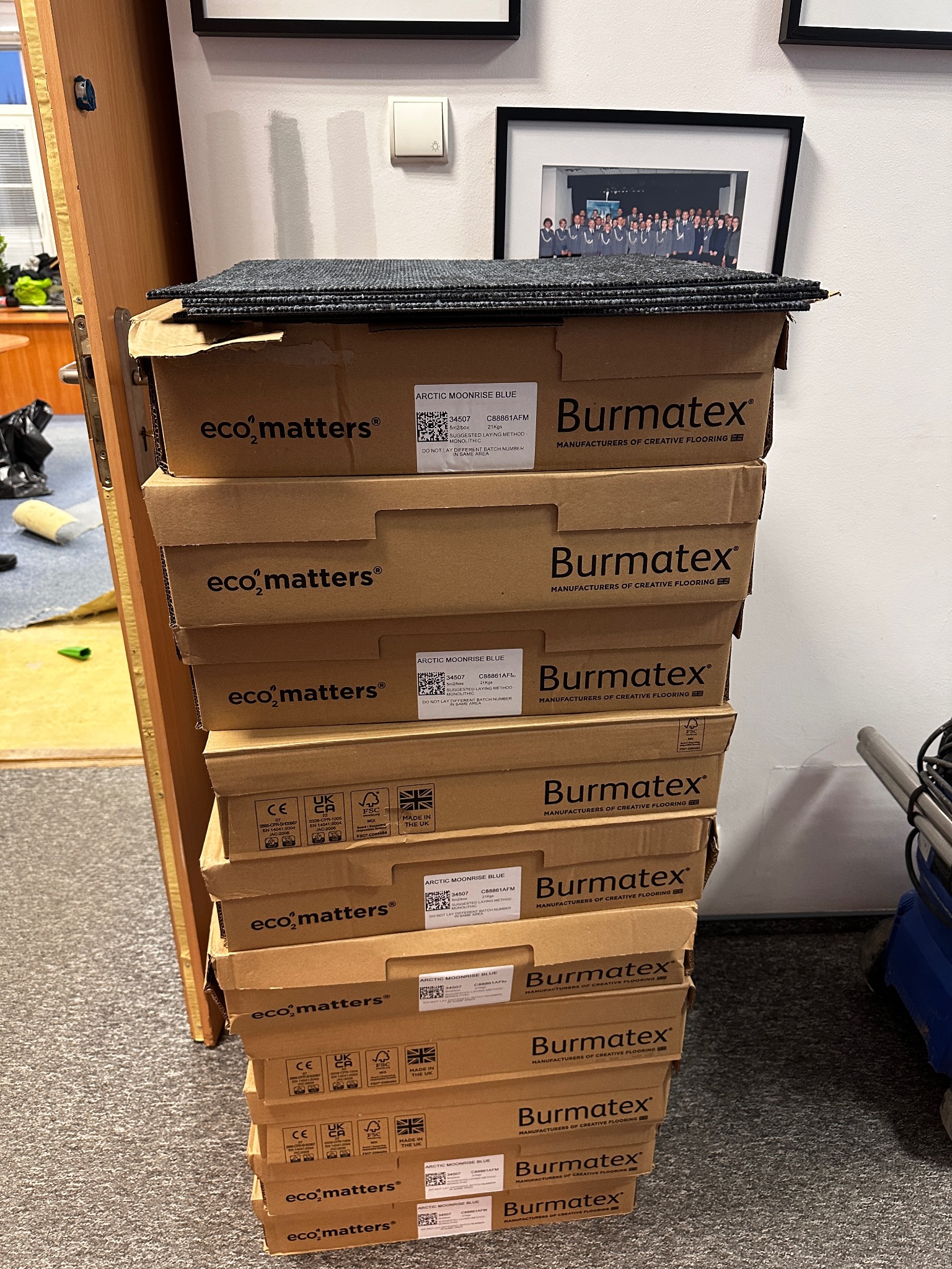 Stos kartonów z szarymi płytkami dywanowymi Burmatex Eco Matters Arctic Moonrise Blue, na wierzchu ułożone płytki, widoczny fragment pomieszczenia z biurkiem i krzesłem.