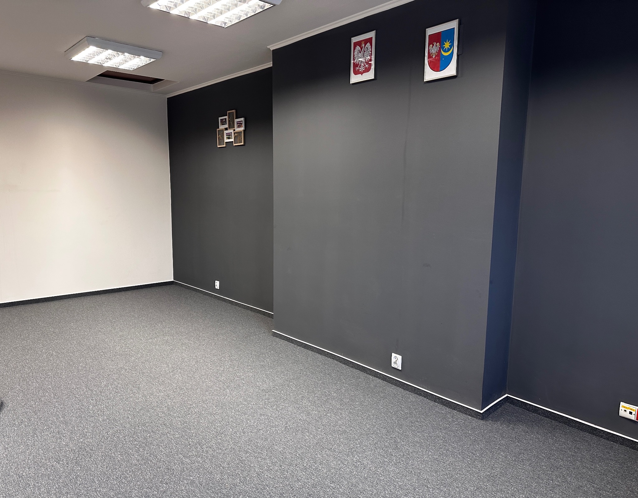 Wnętrze biura po remoncie: szara wykładzina dywanowa, pomalowane ściany w kolorze grafitowym i białym, na ścianach wiszą godła i ramki ze zdjęciami.
