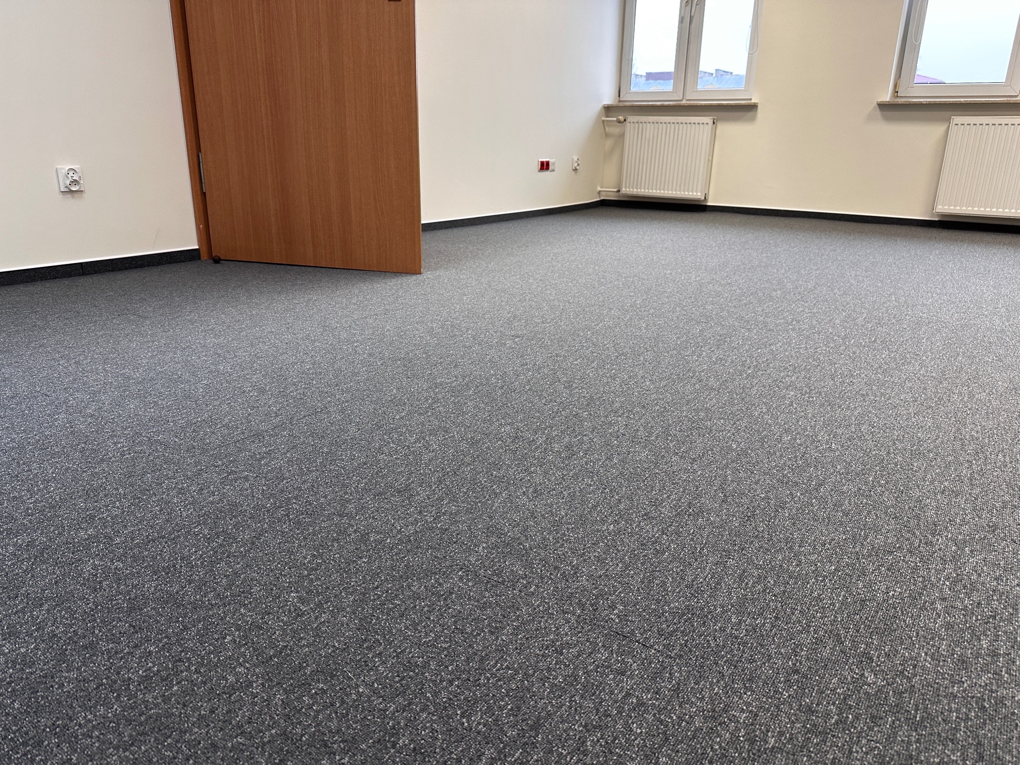 Wnętrze biura z nową, szarą wykładziną dywanową, widoczne grzejniki, drzwi i okna z widokiem na miasto.