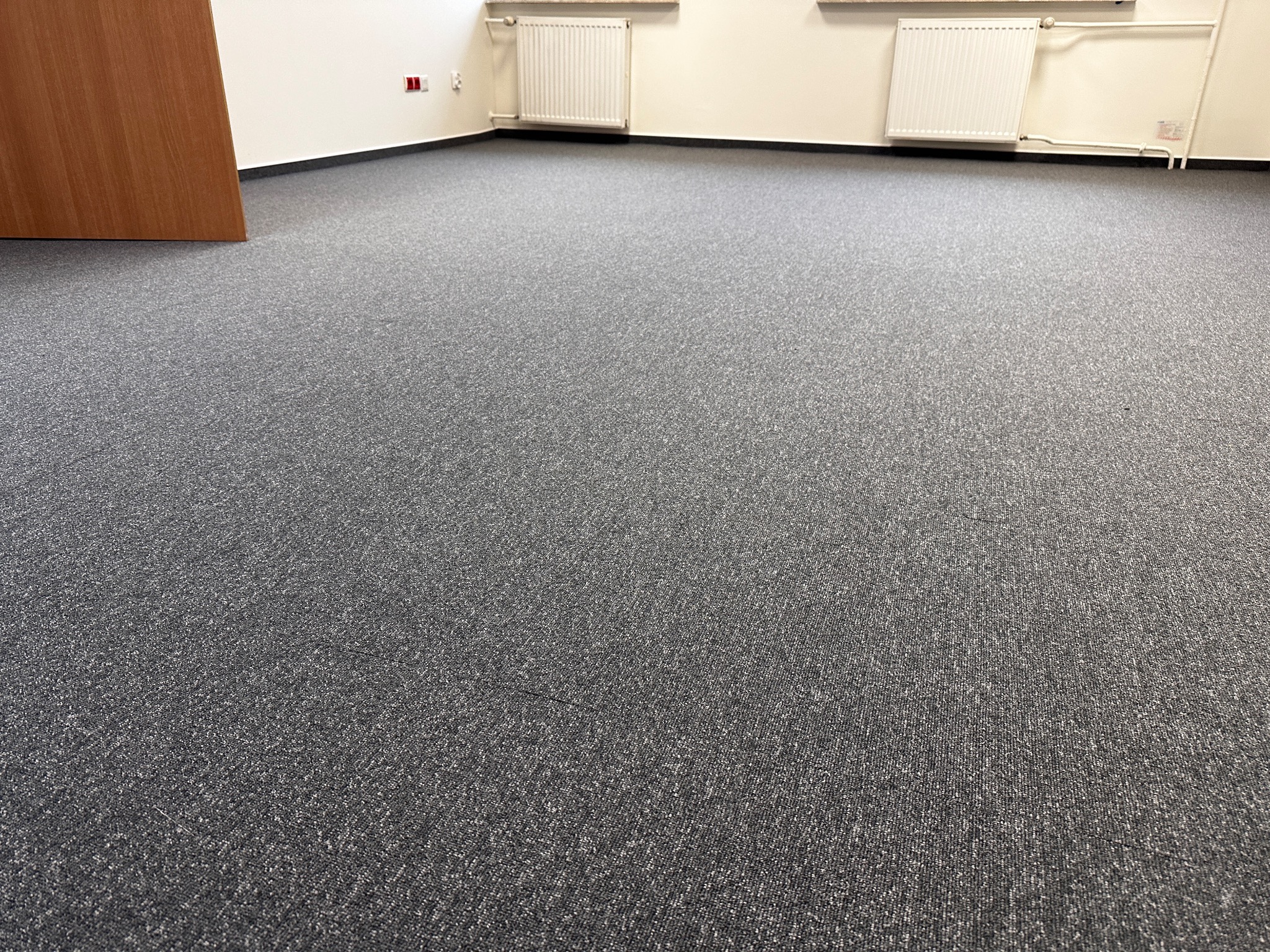 Szara wykładzina dywanowa w biurze, widoczne grzejniki i drzwi, neutralne oświetlenie.