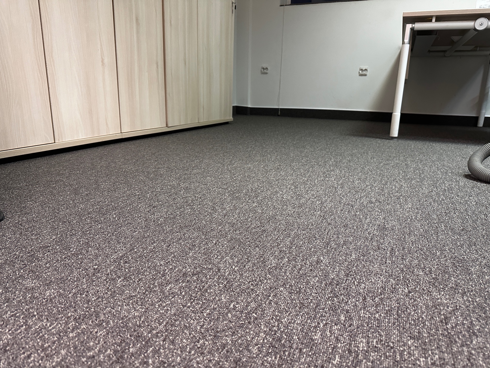 Szara wykładzina dywanowa w biurze, widok z niskiej perspektywy, z fragmentem biurka i szafy w tle.