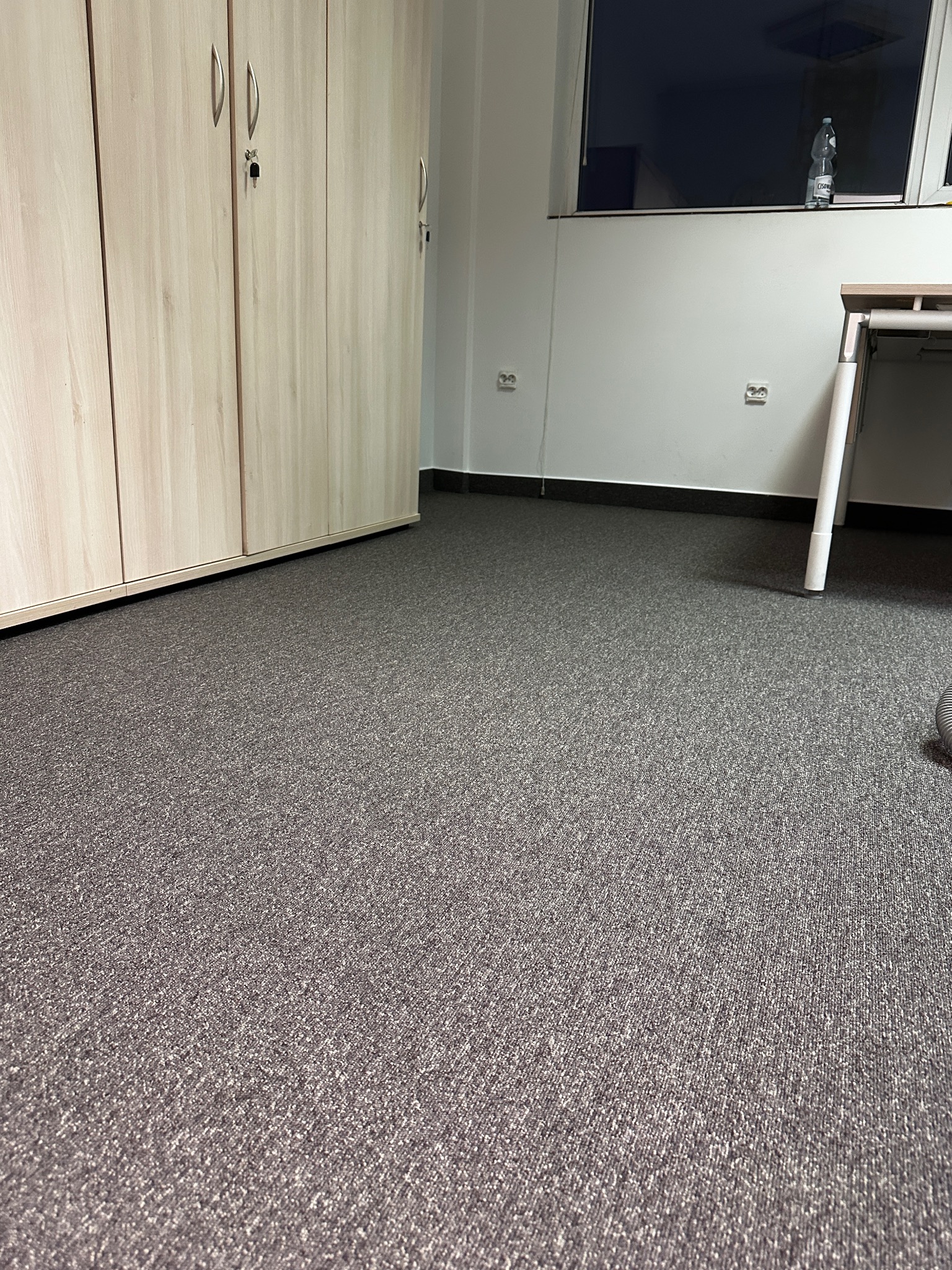 Szara wykładzina dywanowa w biurze, widok na szafę zamykaną na klucz i fragment biurka przy oknie z butelką wody.