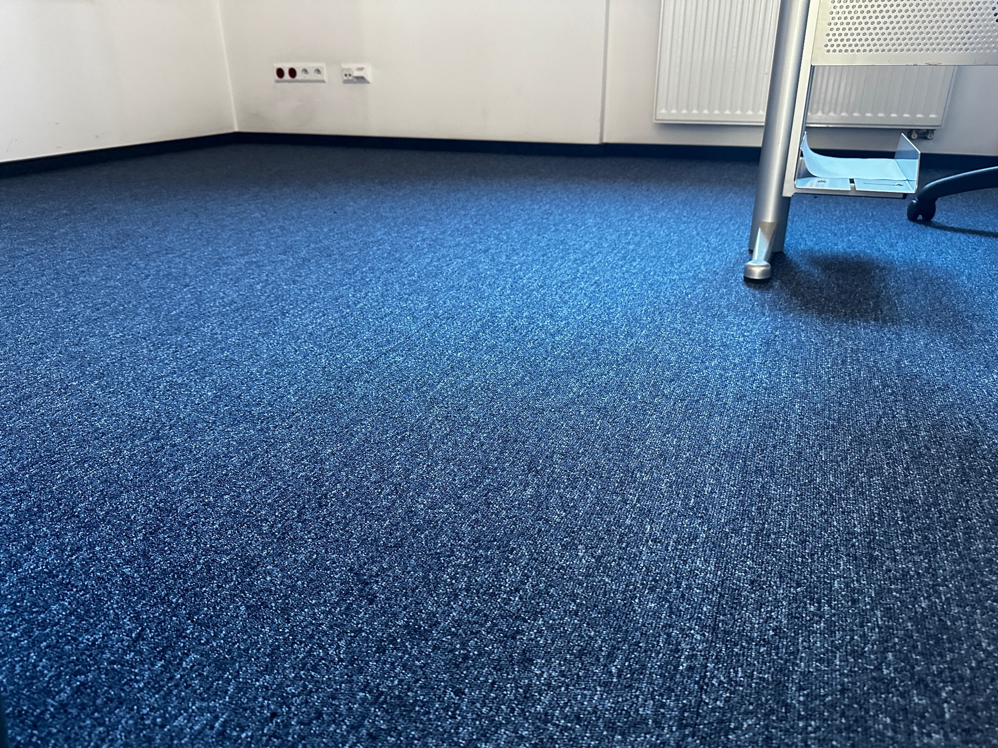 Gładka, granatowa wykładzina dywanowa w biurze, widoczny fragment nogi biurka i gniazdka elektryczne przy ścianie.