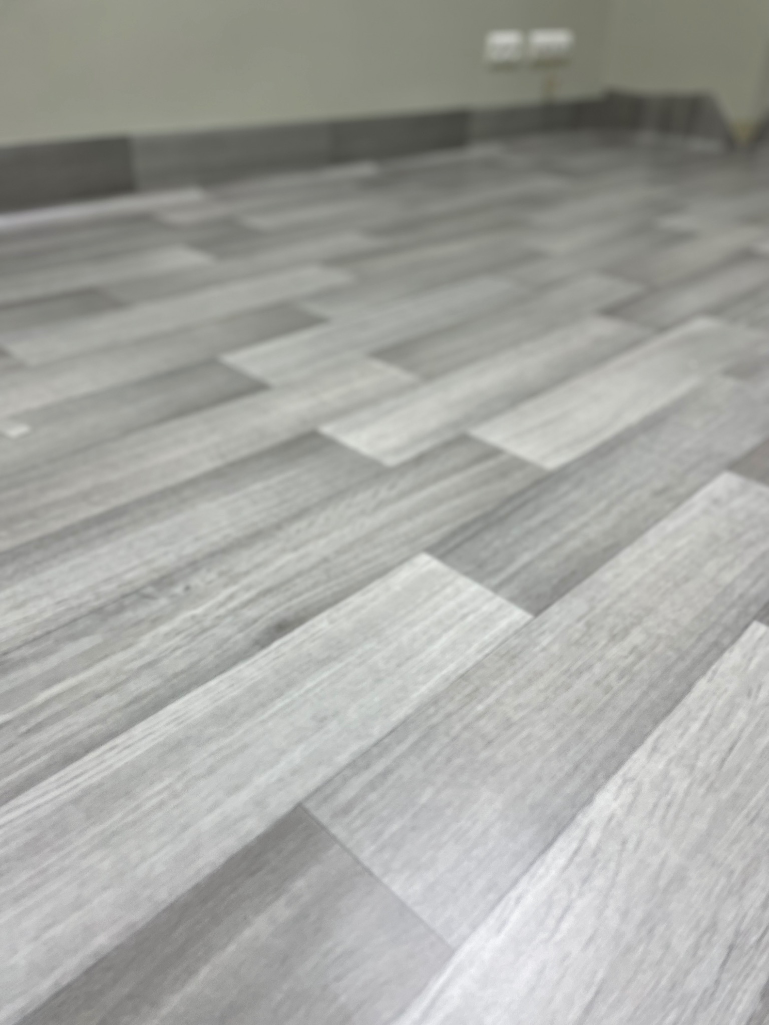 Jasnoszare panele podłogowe ułożone w jodełkę, widok z niskiej perspektywy, z rozmytym tłem ściany i listwy przypodłogowej.