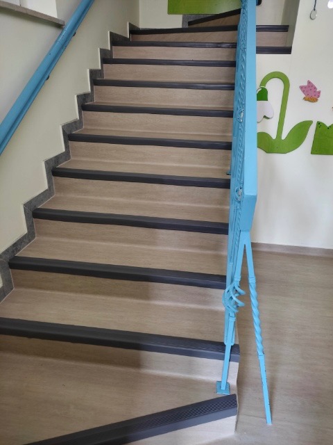 Schody wykończone panelami z czarnymi listwami antypoślizgowymi, z jasnoniebieską metalową balustradą i dekoracjami ściennymi w tle.