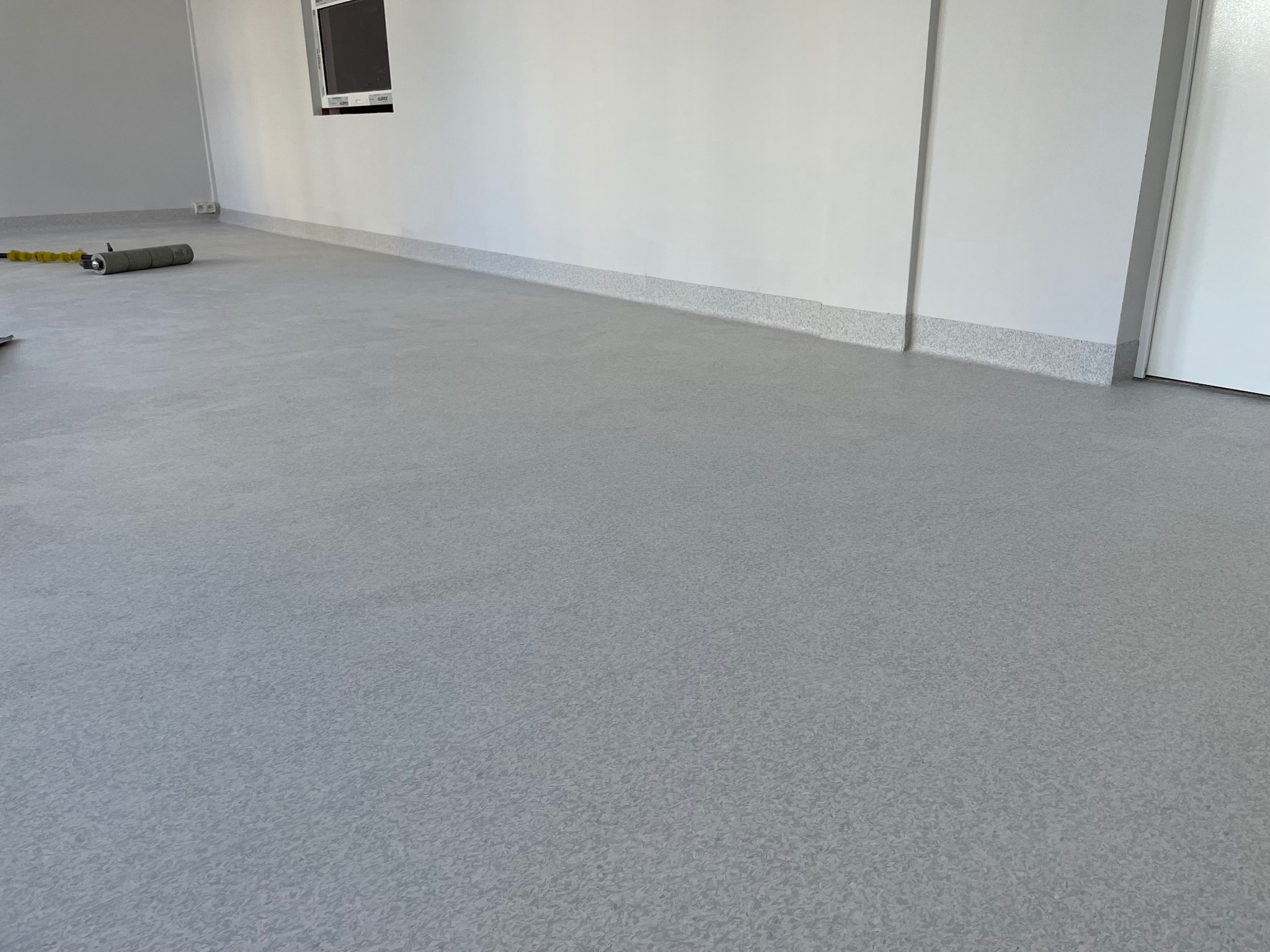 Szara wykładzina podłogowa w jasnym pomieszczeniu biurowym, widoczny wałek dociskowy przy ścianie.