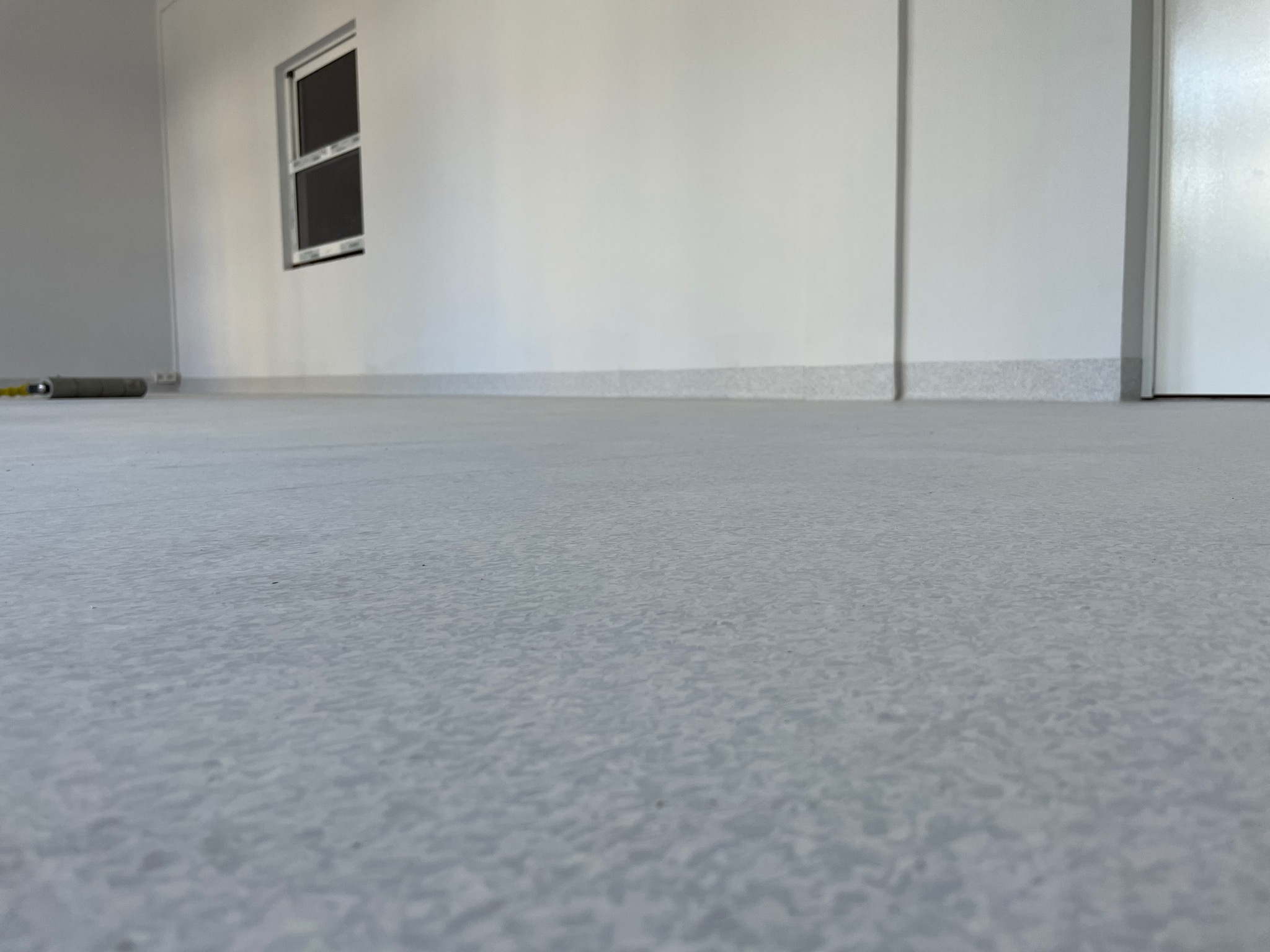 Jasnoszara, wzorzysta podłoga w jasnym pomieszczeniu, widoczne okno i fragment ściany z listwą przypodłogową, wałek malarski leżący przy ścianie.