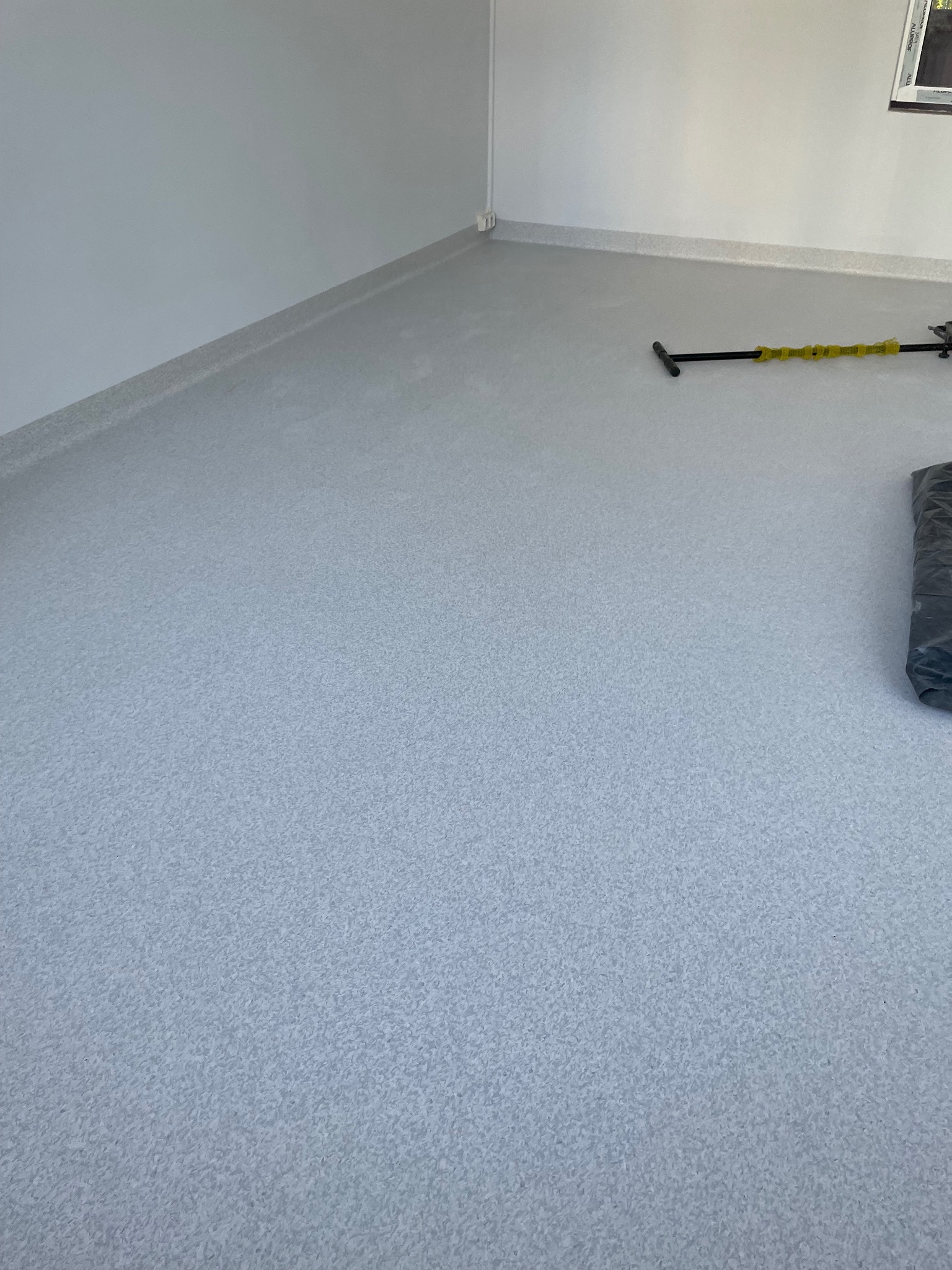 Jasnoszara podłoga z delikatnym wzorem w narożniku pomieszczenia, widoczne listwy przypodłogowe i narzędzie do układania wykładziny z żółtymi elementami.