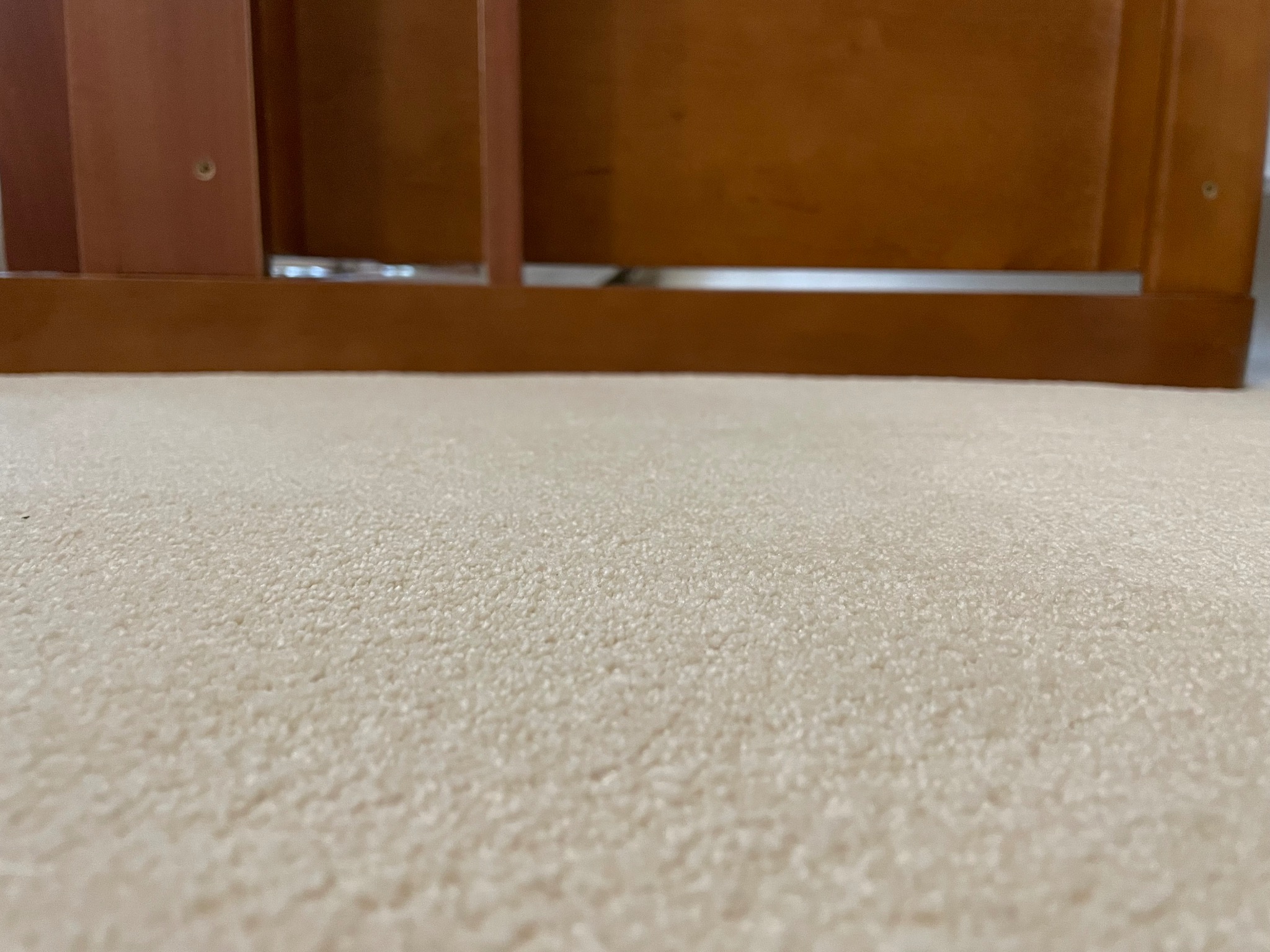 Beżowa, puszysta wykładzina dywanowa z widocznym fragmentem drewnianego mebla w tle.