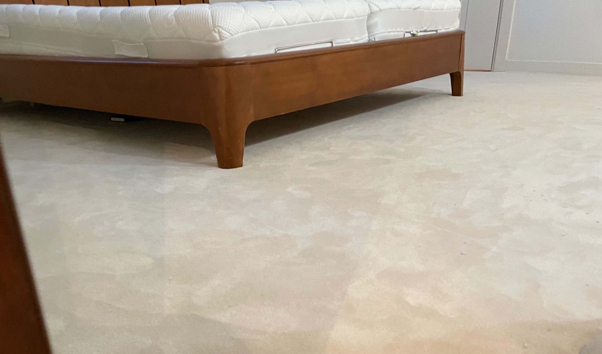 Jasna, kremowa wykładzina dywanowa z widocznym fragmentem drewnianego łóżka z materacem w tle.