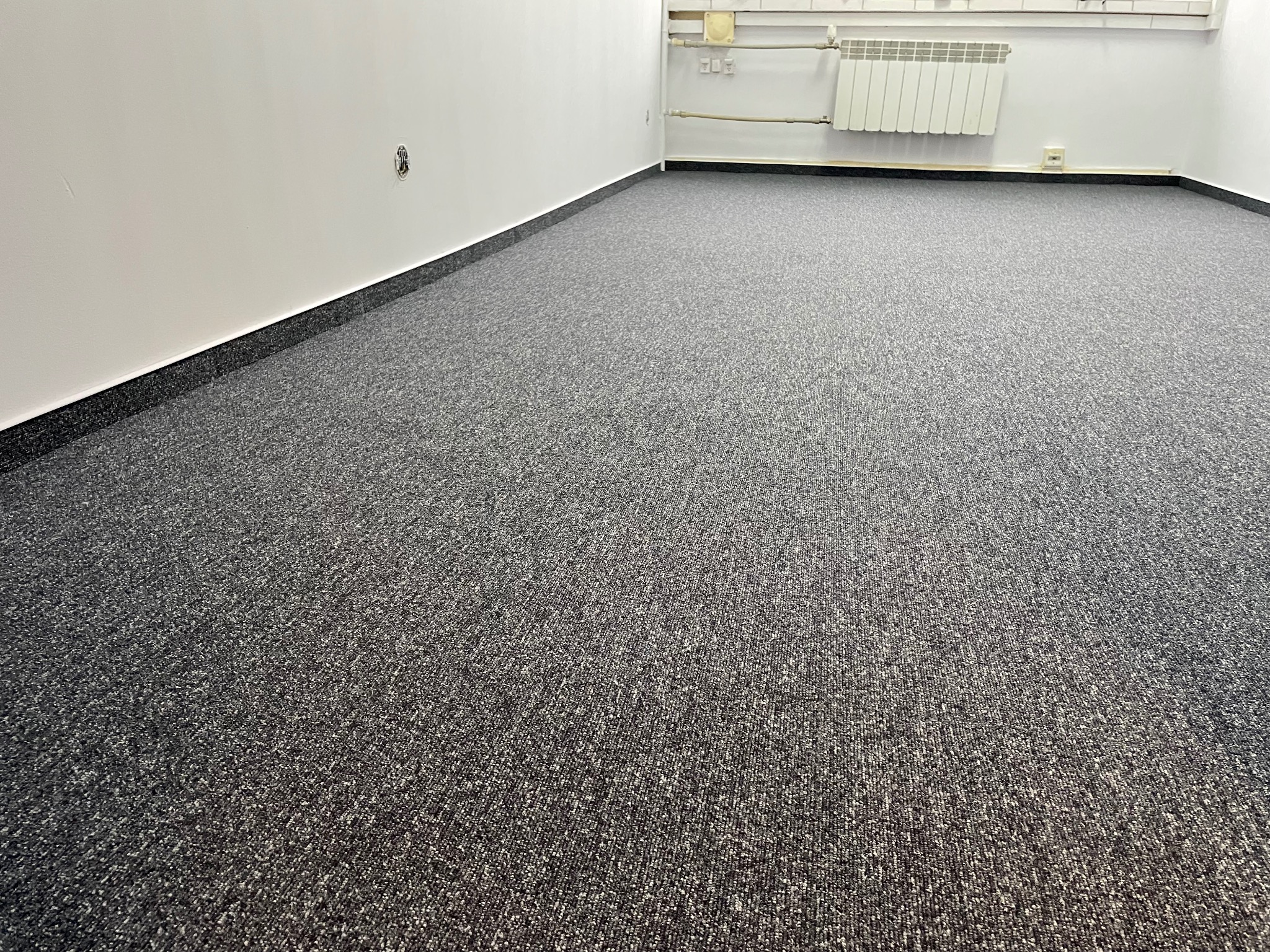Wnętrze biura z nową, szarą wykładziną dywanową o drobnej strukturze, widoczna listwa przypodłogowa i fragment grzejnika na białej ścianie.