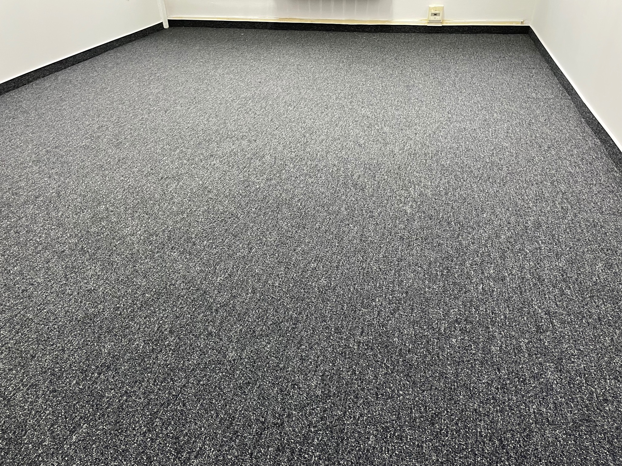 Szara, teksturowana wykładzina dywanowa w pustym pomieszczeniu biurowym z białymi ścianami i czarną listwą przypodłogową.