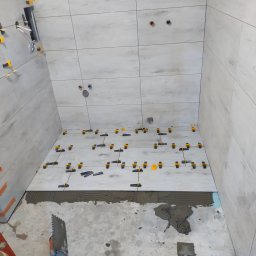 Usługi Remontowe Robert Balcerzak - Świeżo ułożone, jasne płytki podłogowe i ścienne w łazience, z użyciem klipsów poziomujących, widoczne narzędzia glazurnicze i warstwa świeżego kleju. Detal: niebieski fragment izolacji pod płytkami.