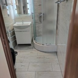 Usługi Remontowe Robert Balcerzak - Wykończona łazienka z kabiną prysznicową, umywalką z szafką, grzejnikiem drabinkowym i podłogą wyłożoną panelami imitującymi drewno, widok zza framugi drzwi.