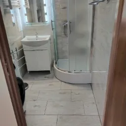 Wykończona łazienka z kabiną prysznicową, umywalką z szafką, grzejnikiem drabinkowym i podłogą wyłożoną panelami imitującymi drewno, widok zza framugi drzwi.