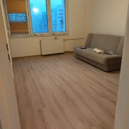 Jasne panele podłogowe ułożone w nowym pokoju z beżowymi ścianami, widoczne okno z żaluzjami i grzejnik, oraz szara sofa z przedmiotami na niej.