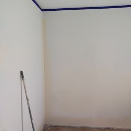 Usługi Remontowe Robert Balcerzak - Pokój w trakcie malowania, z niebieską taśmą malarską przy suficie, wałek oparty o ścianę i kuweta z farbą na podłodze z parkietu. Ściany pomalowane na biało, widoczne ślady po starej farbie.